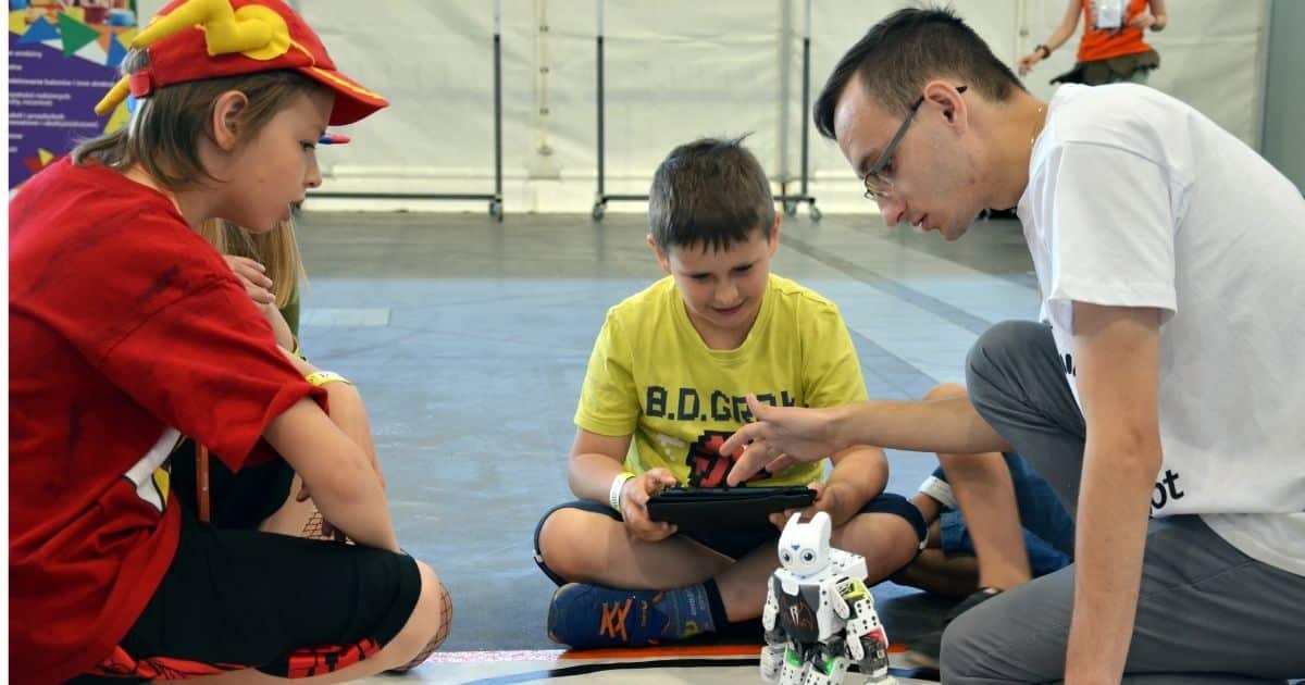 Dwóch chłopców siedzi na podłodze, obok nich klęczy mężczyzna w okularach. Chłopiec w żółtej koszulce trzyma tablet i patrzy na małego robota, który stoi na podłodze. Drugi chłopiec, ubrany w czerwoną koszulkę i czapkę, obserwuje sytuację.