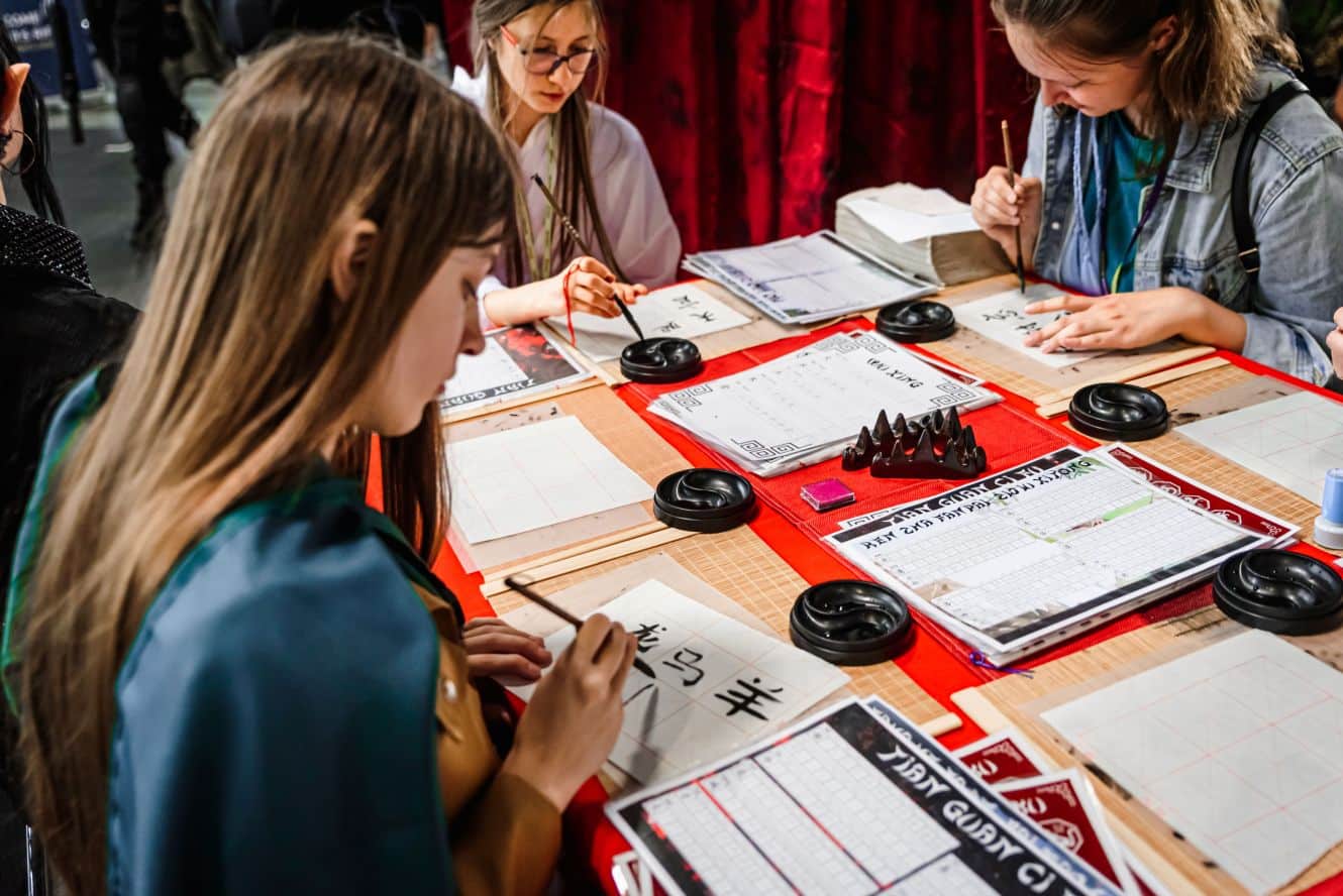 Trzy młode kobiety siedzą przy stole i piszą chińskie znaki pędzelkami na papierze. Na stole leżą czarne naczynia z tuszem, czerwone podkładki i arkusze z szablonami do kaligrafii.