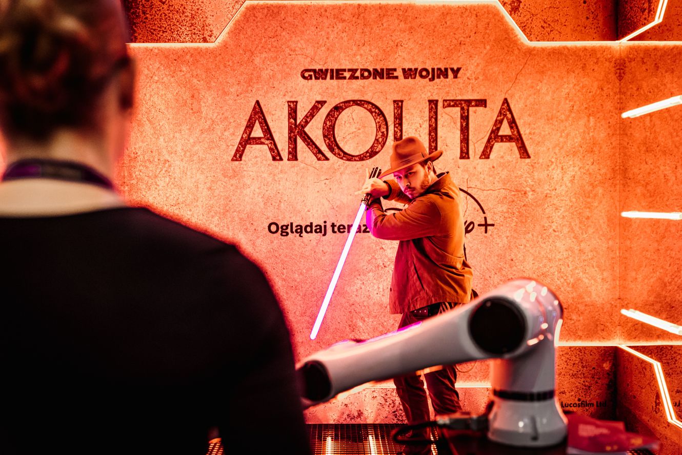Osoba w kapeluszu dzierży różowy miecz świetlny na tle plakatu "Gwiezdne Wojny: Akolita".