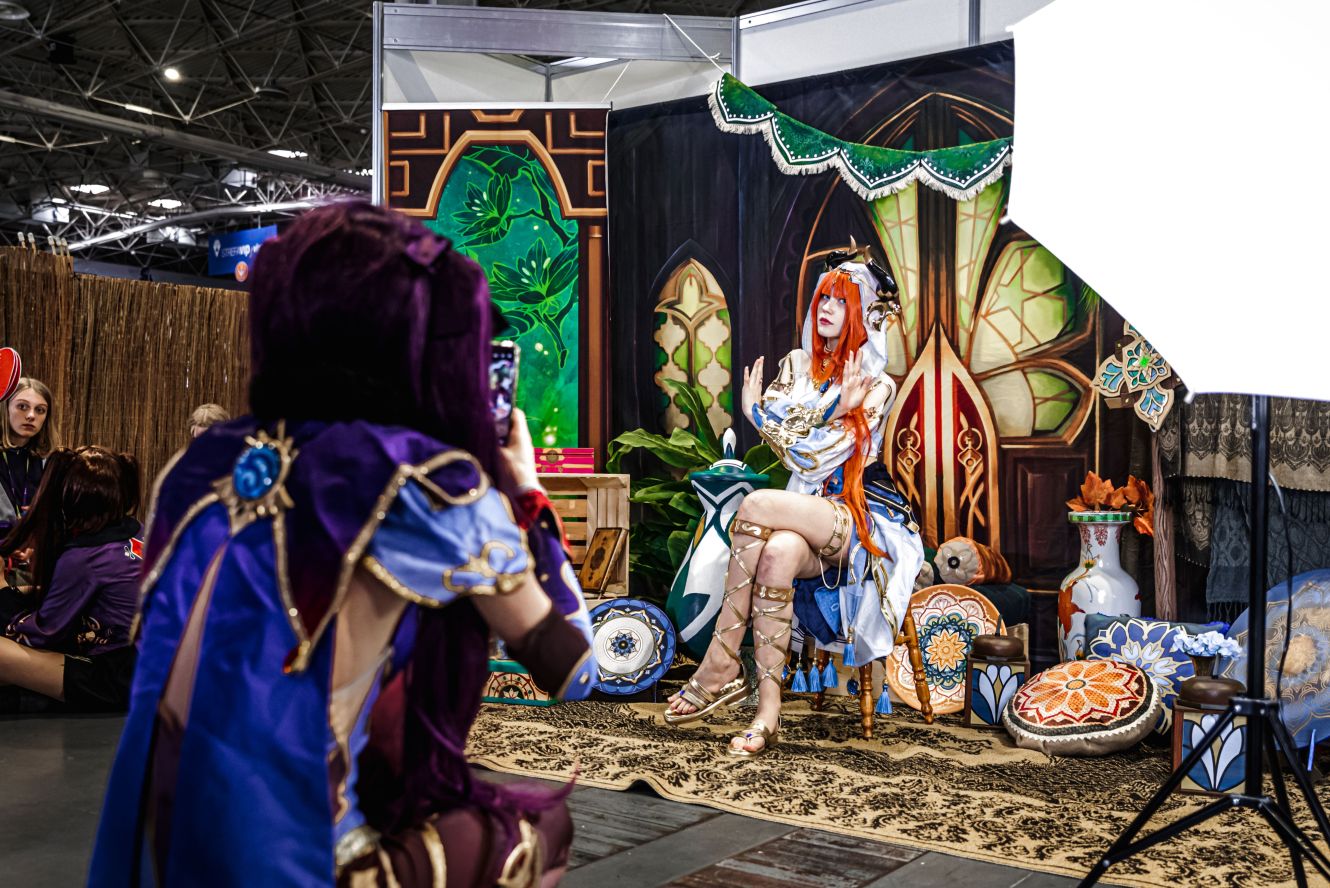 Kobieta przebrana za postać z gry wideo, Nilou z Genshin Impact, siedzi na krześle w stylizowanej scenerii, pozując do zdjęcia robionego przez osobę w kostiumie zza aparatu.