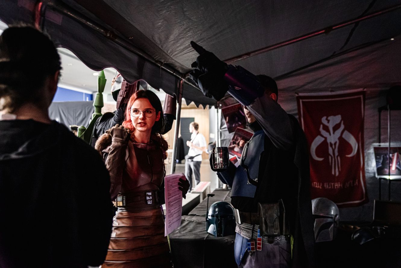 Kobieta w kostiumie inspirowanym Star Wars rozmawia z mężczyzną w kostiumie Mandalorianina przy stoisku z gadżetami, w tle flaga z symbolem czaszki Mythosaurusa.