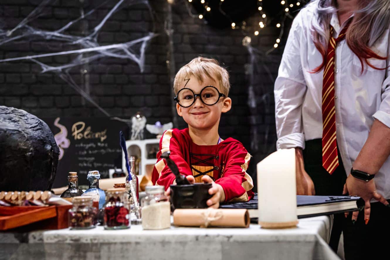 Chłopiec w kostiumie Harry'ego Pottera, z okularami i blizną na czole, miesza składniki w czarnym moździerzu, siedząc przy stole z magicznymi rekwizytami, obok stoi osoba w stroju ucznia Hogwartu, w tle dekoracje halloweenowe.