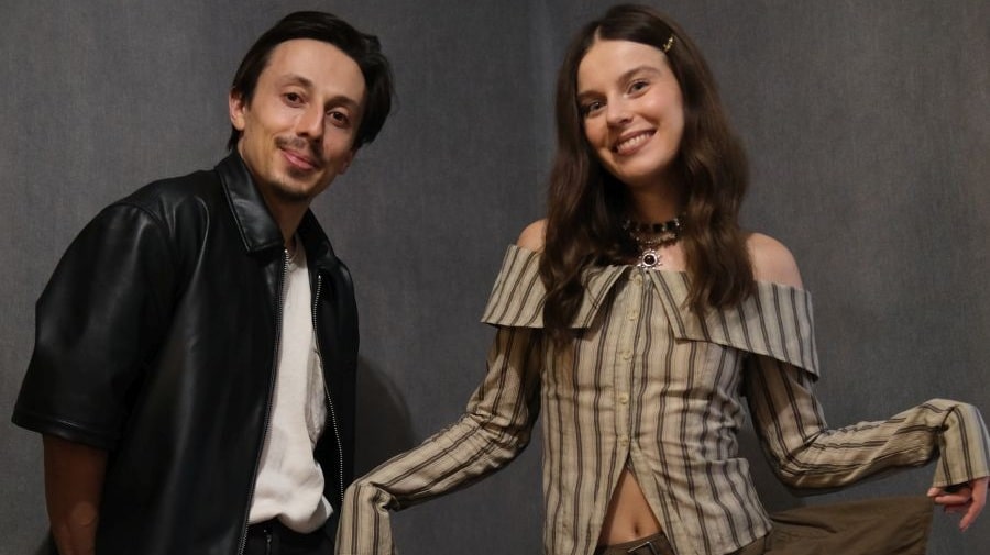 Dwie osoby pozują do zdjęcia na ciemnoszarym, neutralnym tle studyjnym. Po lewej stronie znajduje się osoba o krótkich, ciemnych włosach i lekkim zaroście, ubrana w czarną, skórzaną kamizelkę narzuconą na jasną koszulkę. Jej postura jest swobodna, lekko pochylona w stronę aparatu, a wyraz twarzy spokojny i przyjazny. Po prawej stronie stoi uśmiechnięta osoba z długimi, falującymi brązowymi włosami, częściowo spiętymi ozdobną spinką. Ma na sobie jasną bluzkę w pionowe paski z odkrytymi ramionami i szerokim kołnierzem opadającym na ramiona. Ręce obu osób są rozłożone na boki w lekkim, otwartym geście, co nadaje zdjęciu naturalny, swobodny charakter i sugeruje dobrą atmosferę podczas sesji.