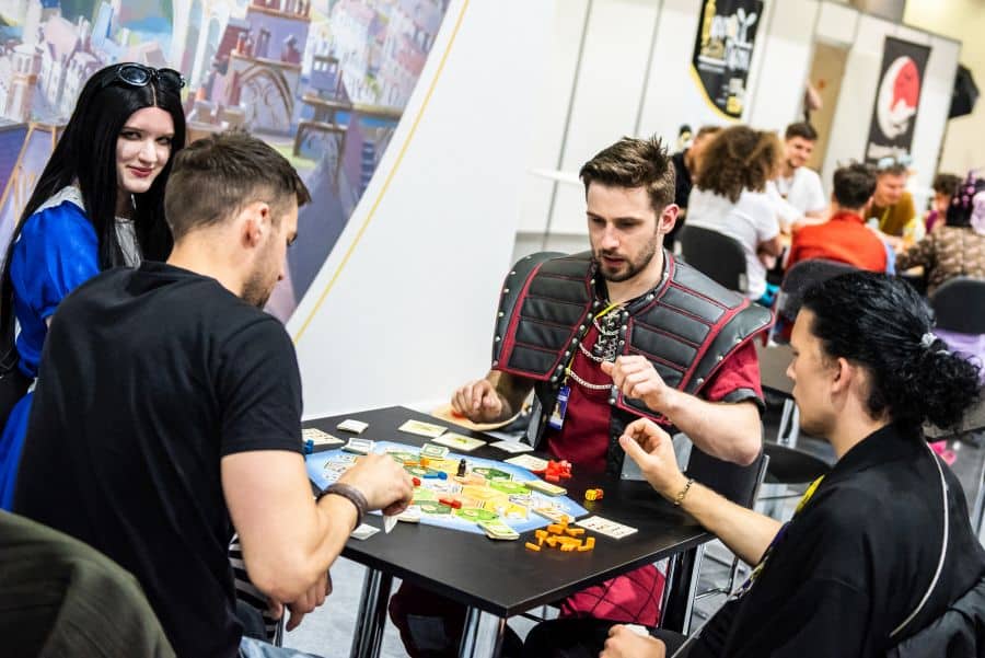 Cztery osoby siedzą przy małym stoliku i grają w kolorową grę planszową z pionkami i kartami. Jedna z osób ma na sobie strój przypominający kostium fantasy lub historyczny pancerz. W tle widać inne osoby siedzące przy stołach oraz plakaty na ścianach, co sugeruje wydarzenie lub spotkanie związane z grami.