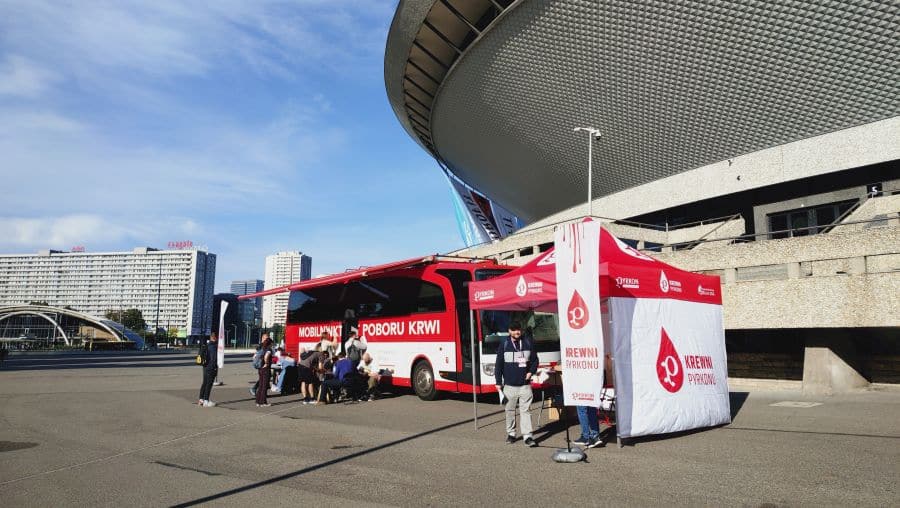 Zdjęcie przedstawia krwiobus Regionalnego Centrum Krwiodawstwa i Krwiolecznictwa w Katowicach. Krwiobus stoi na placu przed katowickim Spodkiem. Przed krwiobusem widać namiot akcji Krewnych Pyrkonu oraz grupę ludzi czekających na oddanie krwi.